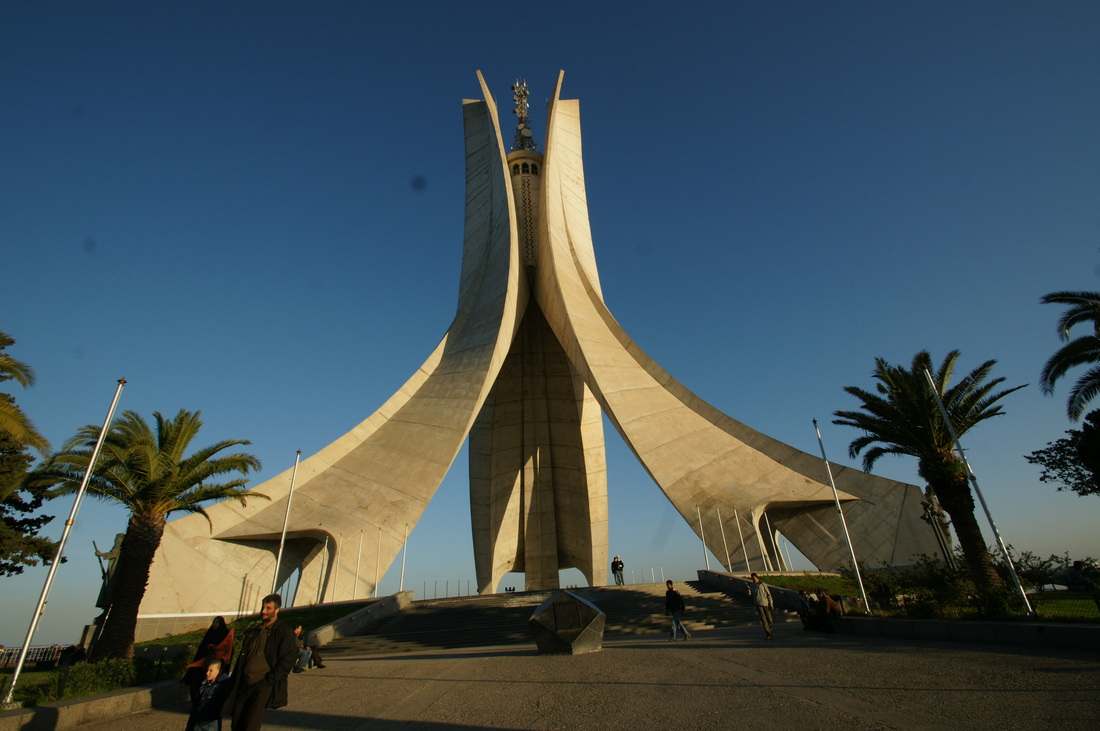 Edifício, construído em forma de três Palmas na Argel. Monumento em uma praça, durante o dia, com pessoas passando, sentadas e fotografando. 