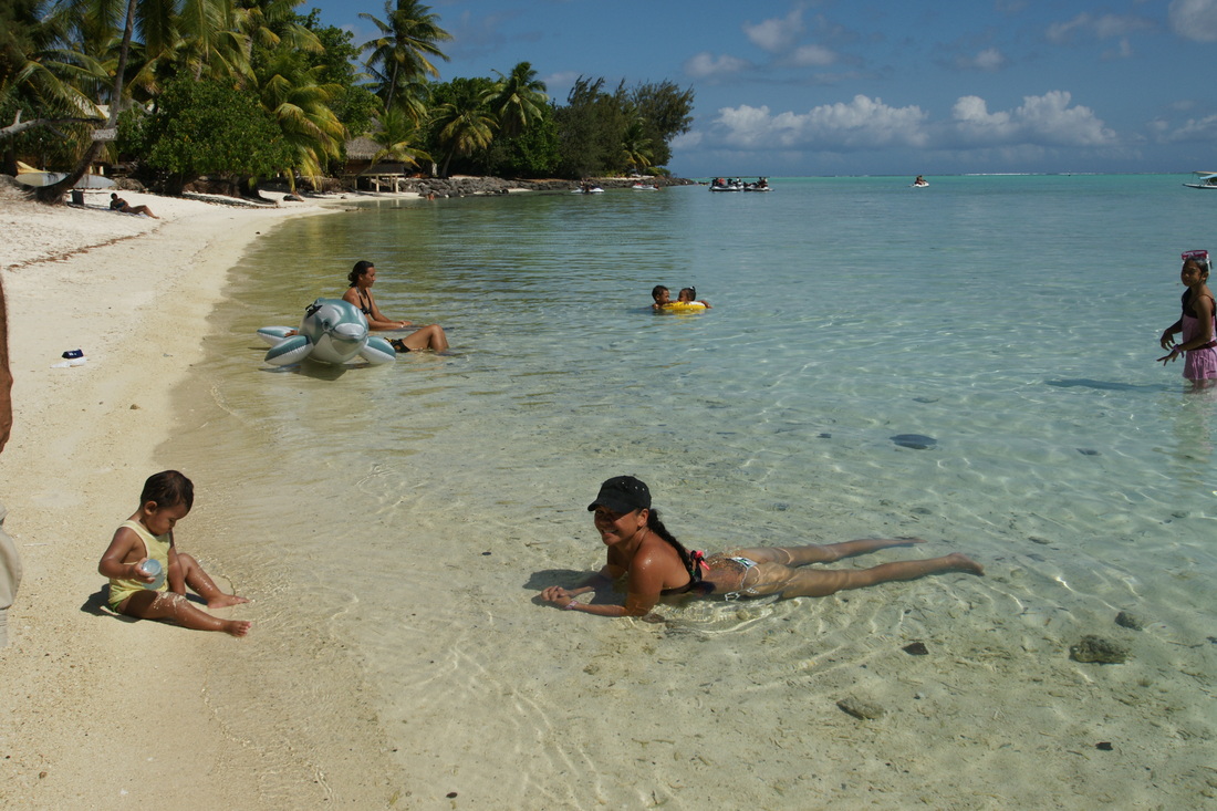 Pessoas em uma praia da Polinésia Francesa, um mar calmo, sem ondas e crianças na água.