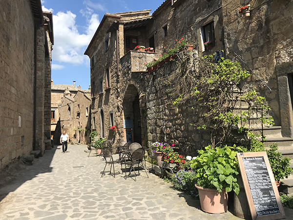CIVITA DI BAGNOREGIO - ITÁLIA, vila rústica com escada, paredes se pedras, vasos com plantas e um homem passando pelo rua.
