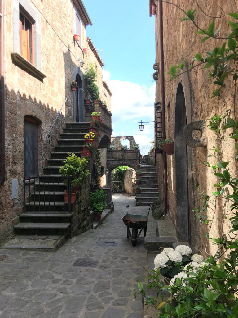 Pequena vila em Civita Di Bagnoregio na Itália, com flores, escadas, chão de pedras, um carrinho, casas com paredes rústicas e vasos de flores nas escadas e paredes.